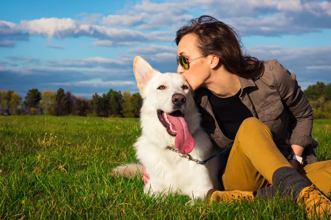 A woman and her dog sitting on the grass and the woman is kissing her dog's forehead