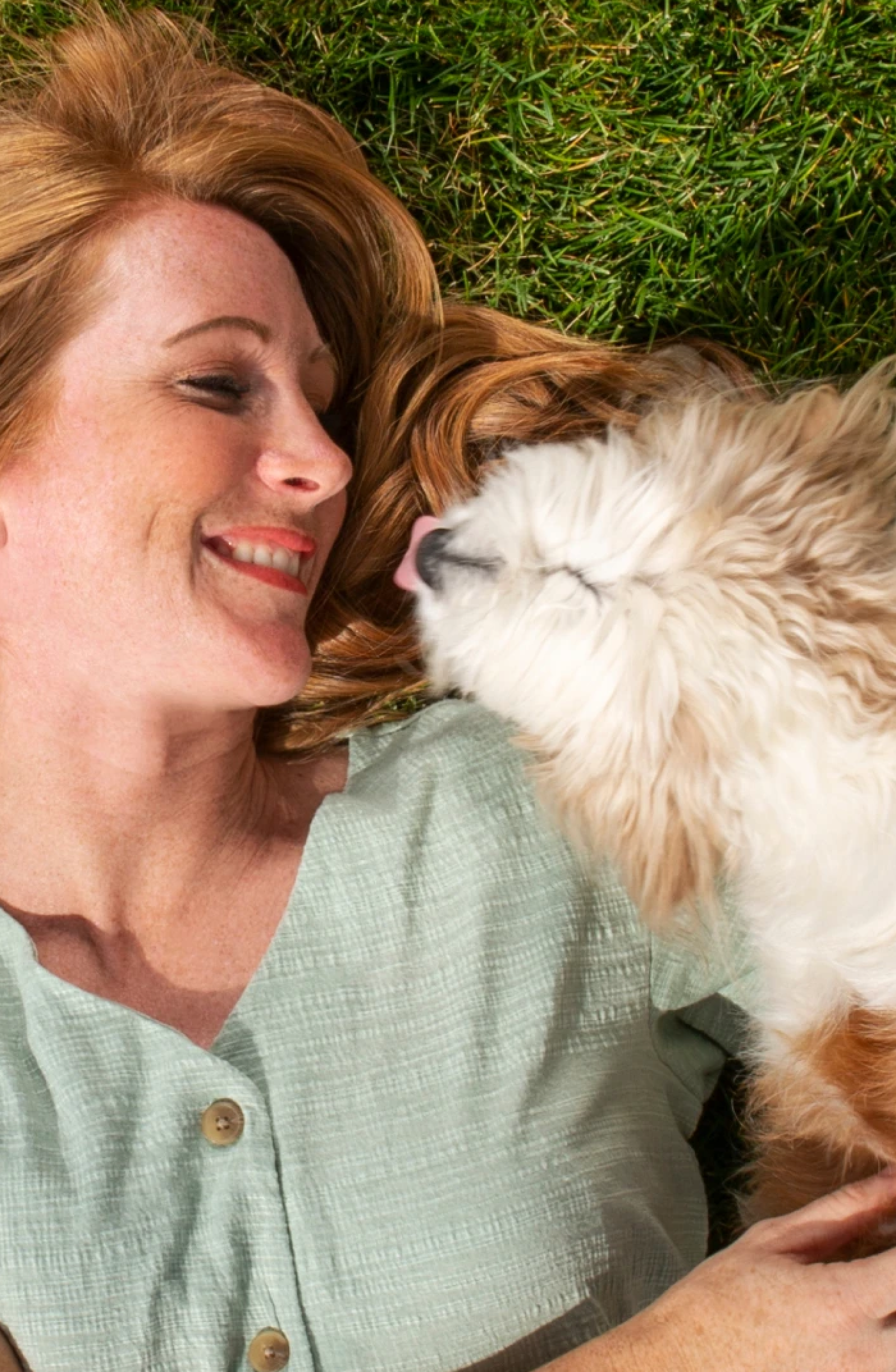 A woman laying on the grass smiling at her dog.